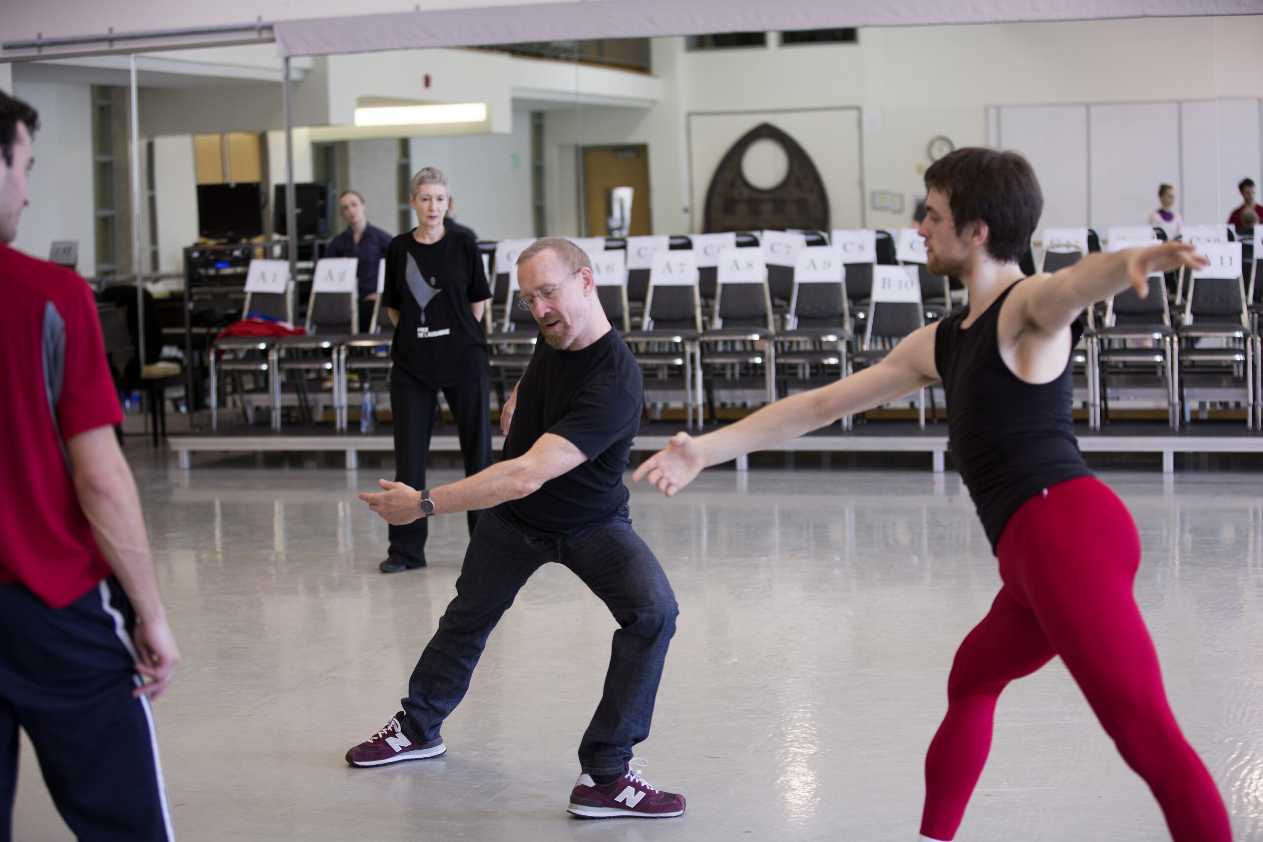 William Forsythe demonstrates a deep lunge, his upper body twisting to his open hip. A dancer facing him imitates the pose, while others stand and observe.