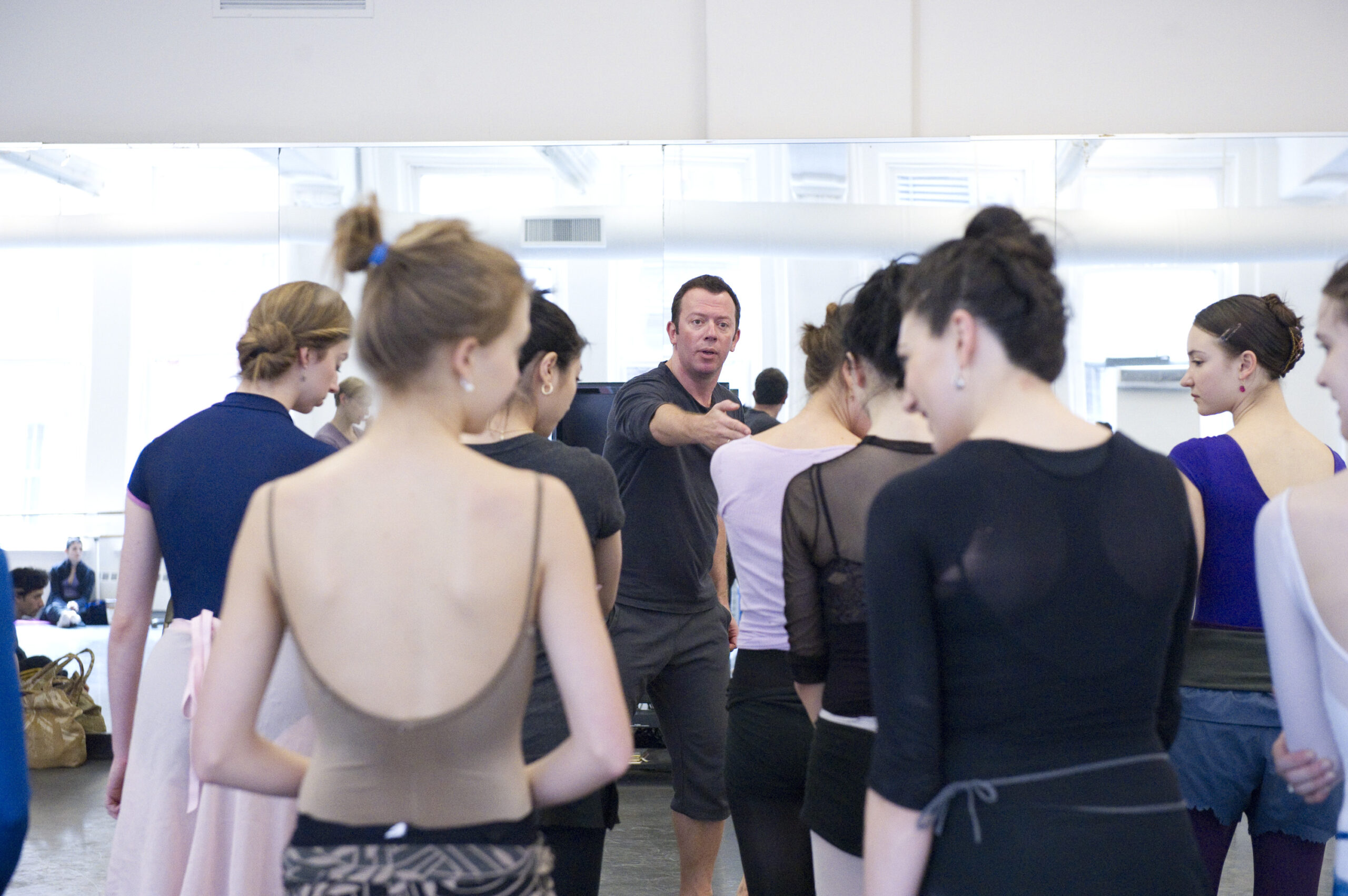 Alexei Ratmansky stands at the front of a mirrored studio, gesturing with one arm as he speaks. Two lines of women in rehearsal clothes stand between him and the camera, their backs to the viewer.