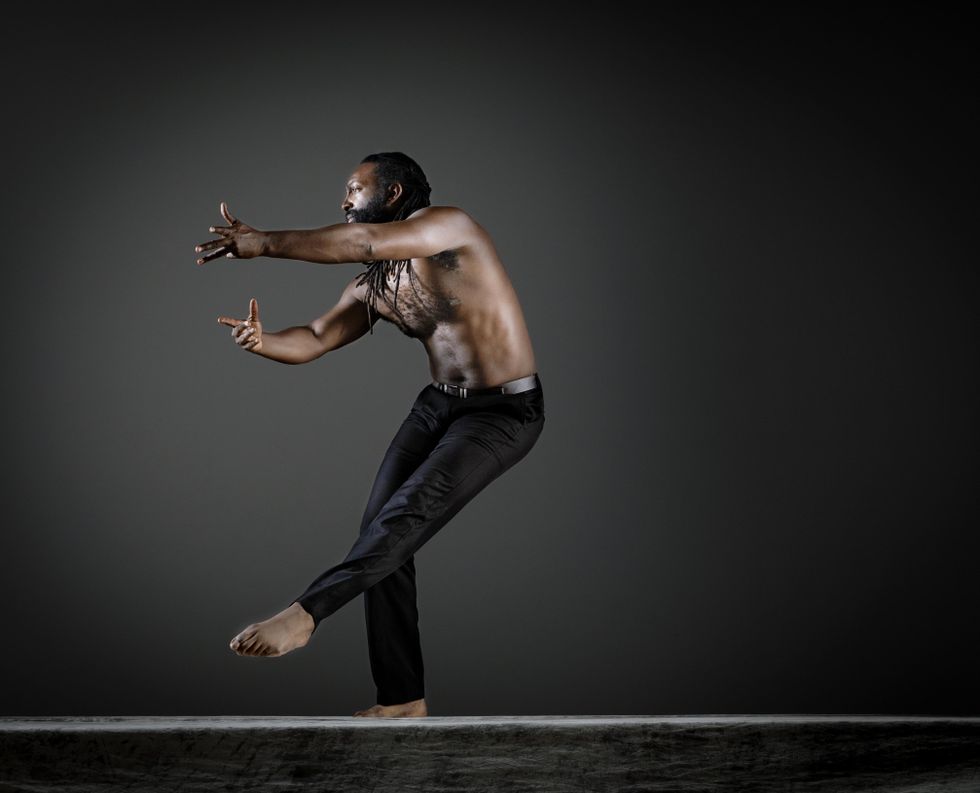 Antoine Hunter reaches to his right side, left leg lifted low and crossed in front of him. He's on a dark stage wearing black pants and no shirt.