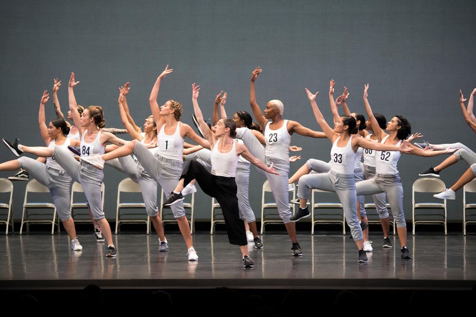 A dozen or so dancers with numbers on their chests lift one leg in high attitude side, behind Monica Bill Barnes who makes the same movement