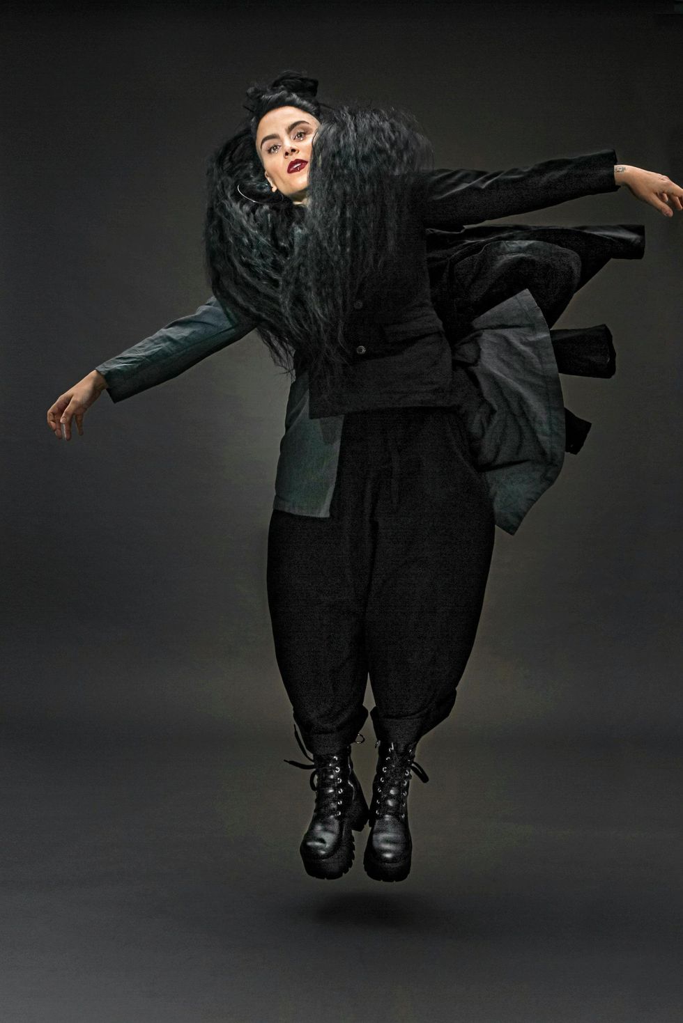 Sonya Tayeh jumps just off the floor, feet together, wearing layers of black and grey which float up around her, following the lines of her arms also floating out to the sides