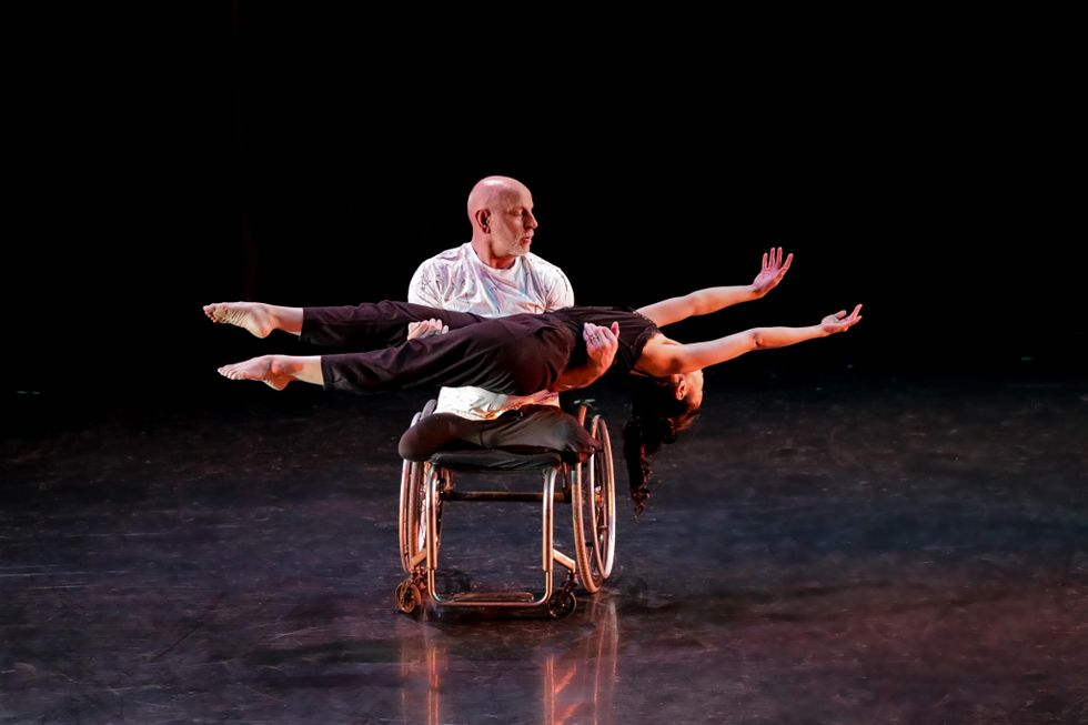 Kris Lenzo in a wheelchair holds Mei-Kuang Chen parallel to the floor, her feet pointed and arms stretched long above her head. He looks down at her.