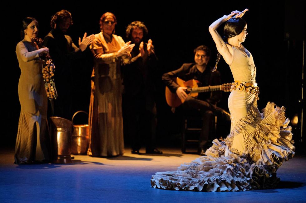 Olga Pericet raises her arms directly overhead, back arched, a long train on her dress flying out below her as musicians in the background perform