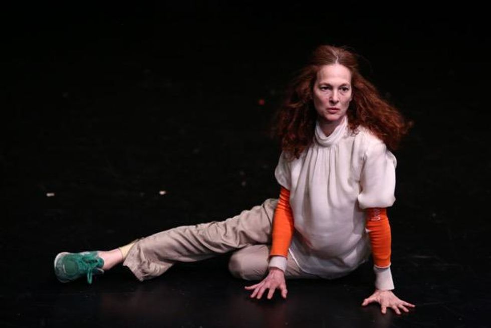 Jodi Melnick sits on one hip on a black stage, her other leg reaching out straight to the side, her splayed hands balancing her
