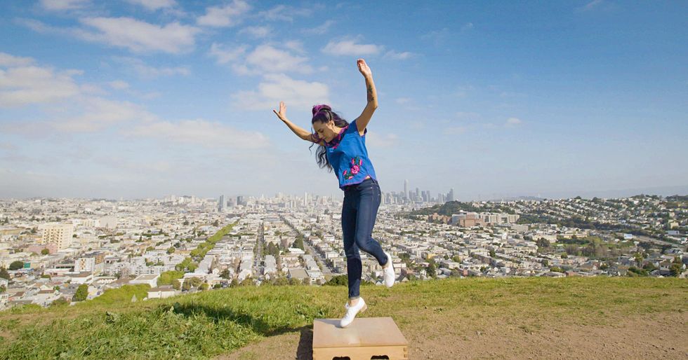 Vanessa Sanchez taps on a small wooden platform on a hill overlooking a city below. One toe touches the platform with the other in the air, hands raised above her head