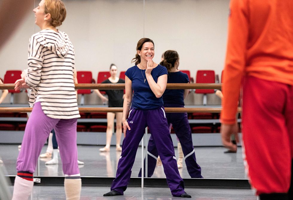 Pam Tanowitz stands in a wide second position in front of a barre and mirror, smiling at dancers in front of her
