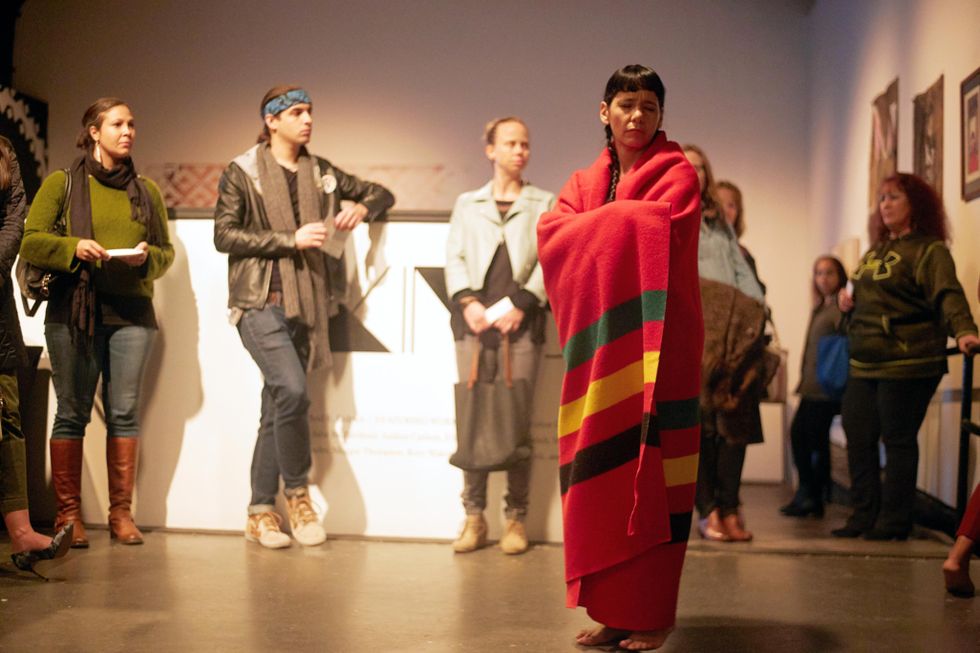 Rosy Simas wraps herself in a long red fabric with multi-colored stripes, eyes closed, as audience members surround her in a gallery environment