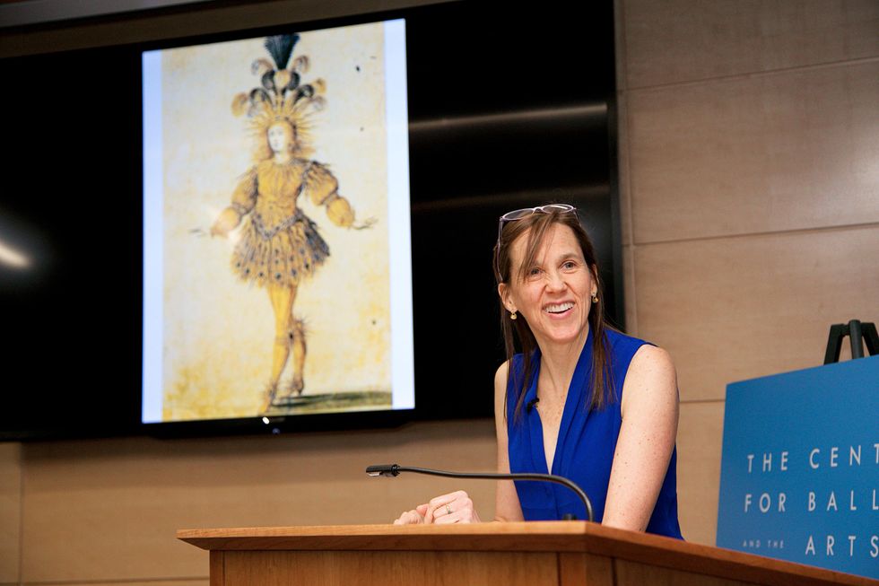Jennifer Homans stands smiling at a lectern, glasses raised on top of her head, a drawing on a screen behind her