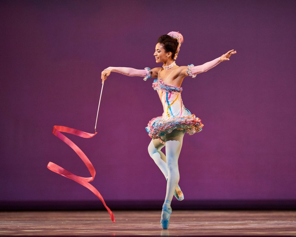 Black ballet dancer Kimberly Marie Olivier stands on pointe with the other foot in coupu00e9, swirling a pink ribbon. 