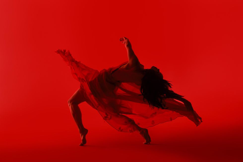 A dancer in a diaphanous skirt hinges toward the ground, legs in a wide fourth and back parallel to the floor. The backdrop and dancer are washed in a vivid red.