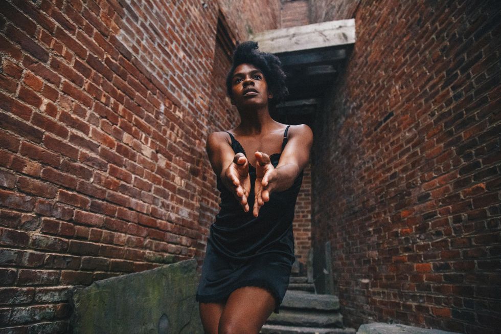 Kayla Farrish gazes above the camera, her arms pressing together in front of her as though offering her nearly closed palms to the camera. Brick walls encroach around her.