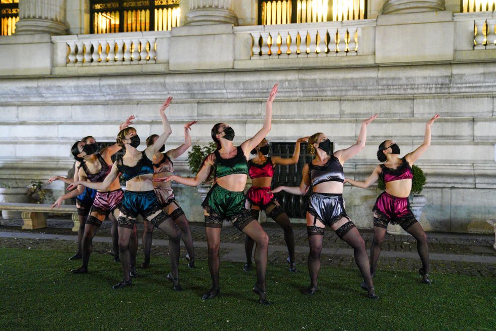 Eight female dancers wearing face masks, silky bra tops and voluminous shorts, and black stockings pose before a white stone building. They gaze at their upraised hands, sitting into their hips.