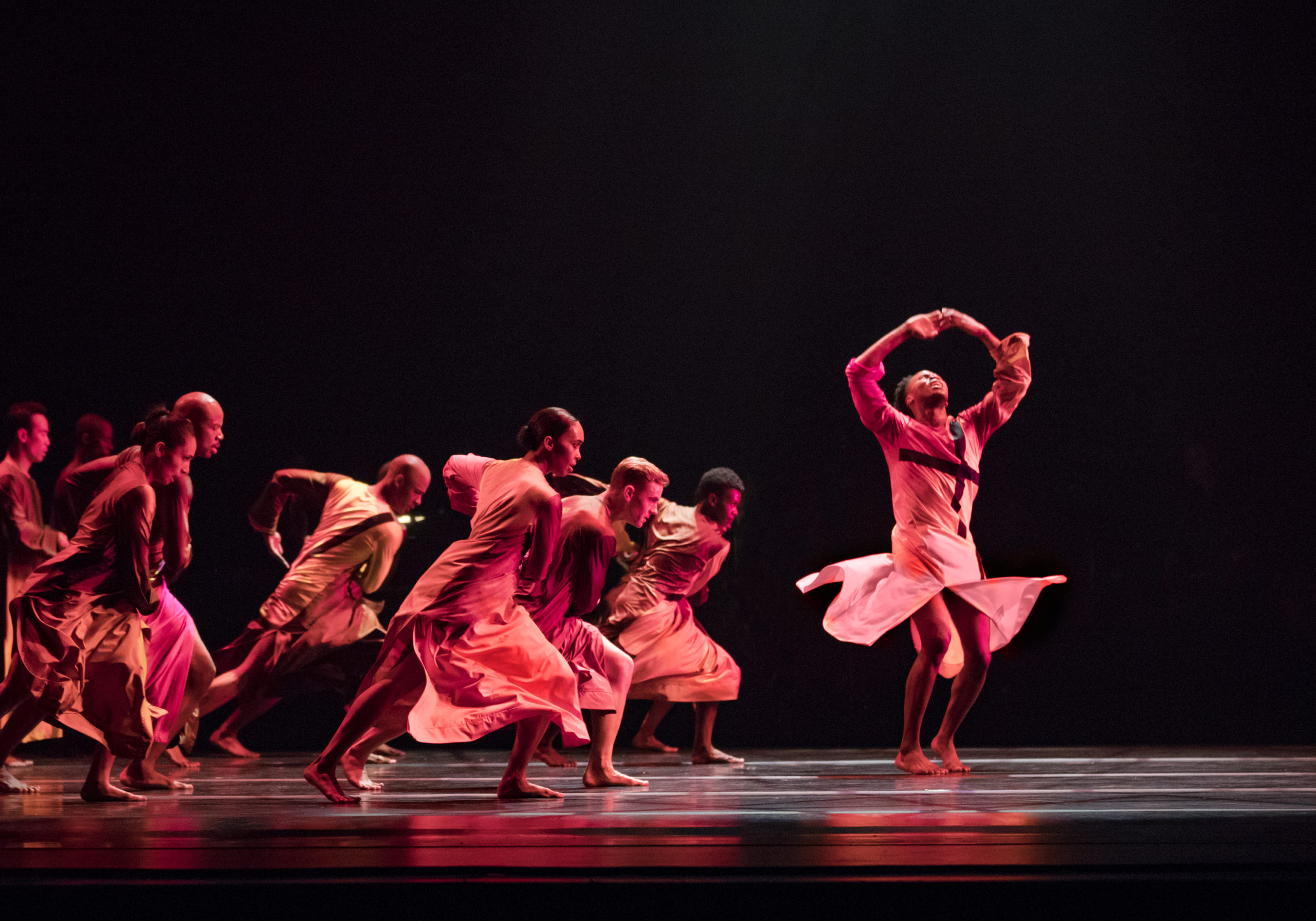 A group of dancers lean forward in a deep lunge, facing a soloist spinning with his hands above his head, looking up