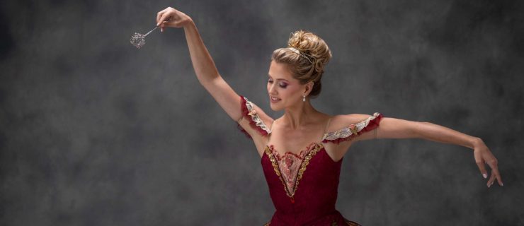 Against a hazy grey backdrop, a smiling dancer poses in a crimson classical tutu with gold trim. Her right arm is raised and points a delicate wand down and out; her gaze follows it. Her left arm is extended in second position. She wears a delicate tiara that frames her voluminous blonde bun.