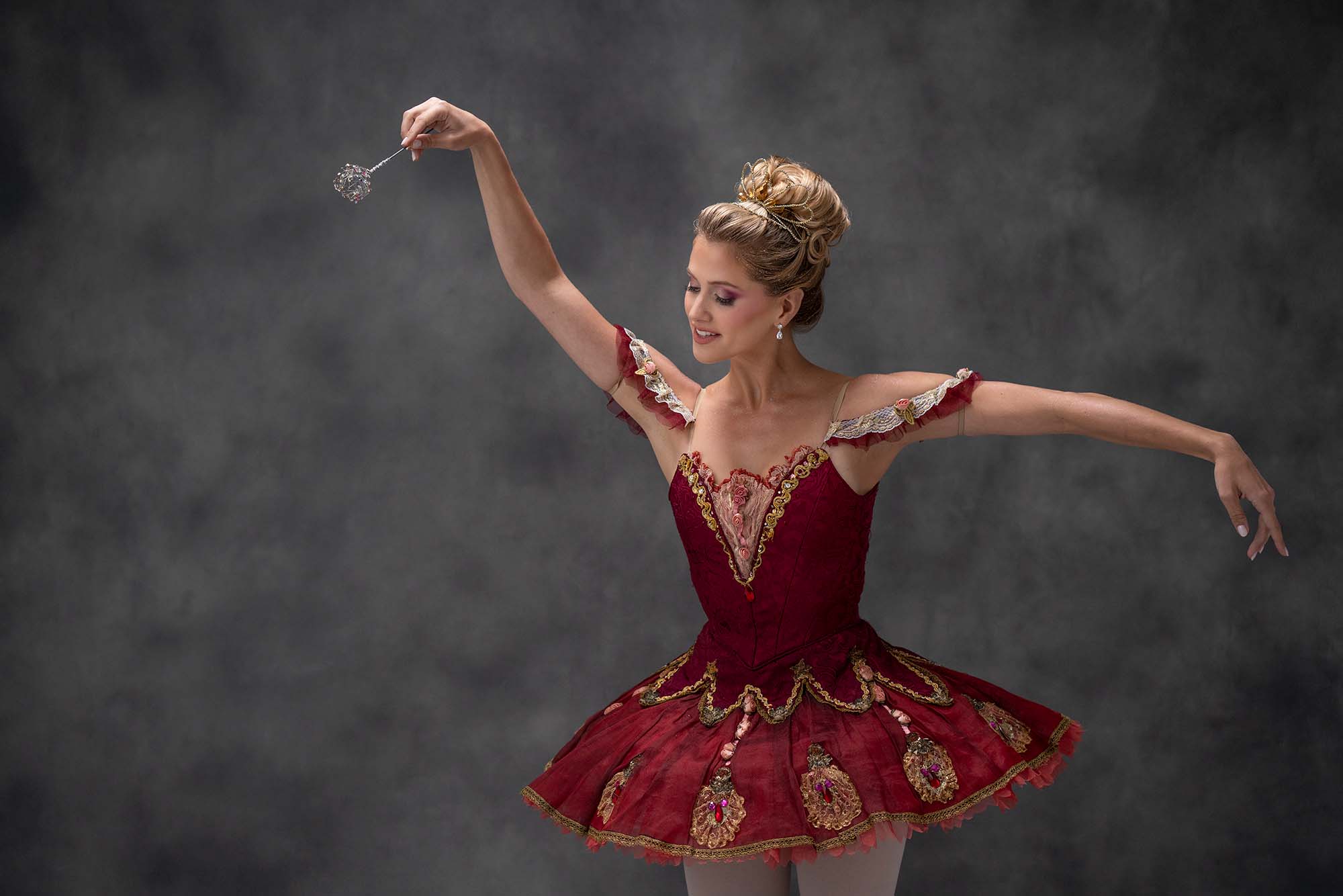 Against a hazy grey backdrop, a smiling dancer poses in a crimson classical tutu with gold trim. Her right arm is raised and points a delicate wand down and out; her gaze follows it. Her left arm is extended in second position. She wears a delicate tiara that frames her voluminous blonde bun.