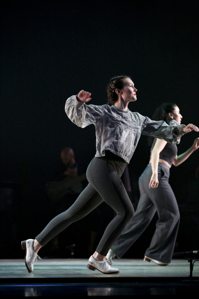 A white female tap dancer lunges forward, her arms out to the sides for balance, in front of another dancer