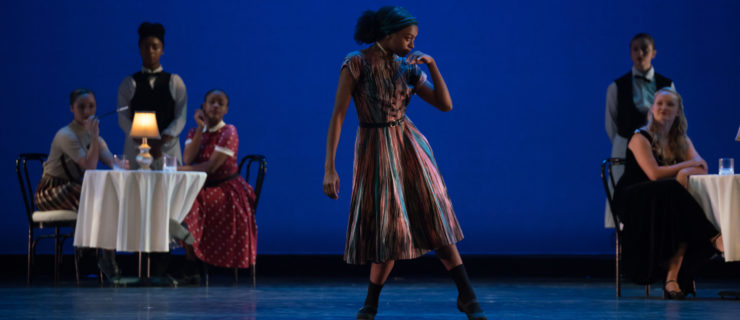 On a blue-lit stage, a woman in a multi-colored striped dress rolls through her feet, hand raised to her mouth as she looks darkly to her left. Upstage, dancers sit watching from two round dinner tables; two others stand in the guise of waiters.