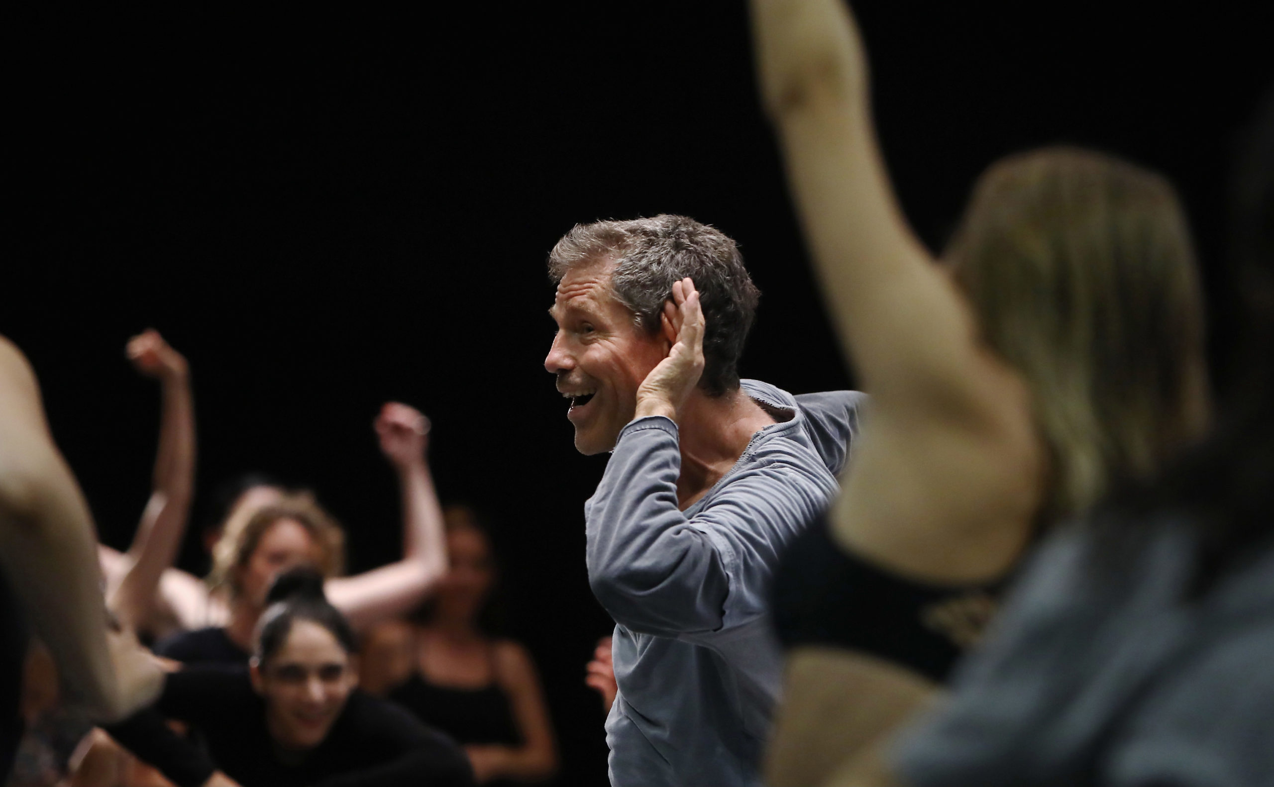 Ohad Naharin holds a hand to his ear, mouth open, surrounded by dancing bodies