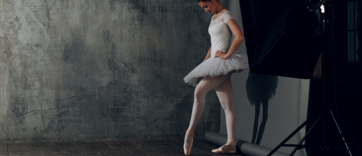 A ballerina in a tutu stands backstage looking down at one pointed foot