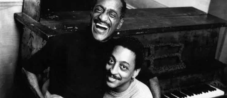 In a black and white archival photo, Sammy Davis Jr. stands beside Gregory Hines, who is seated at a piano bench. Davis Jr. laugh and Hines smiles as the two embrace.