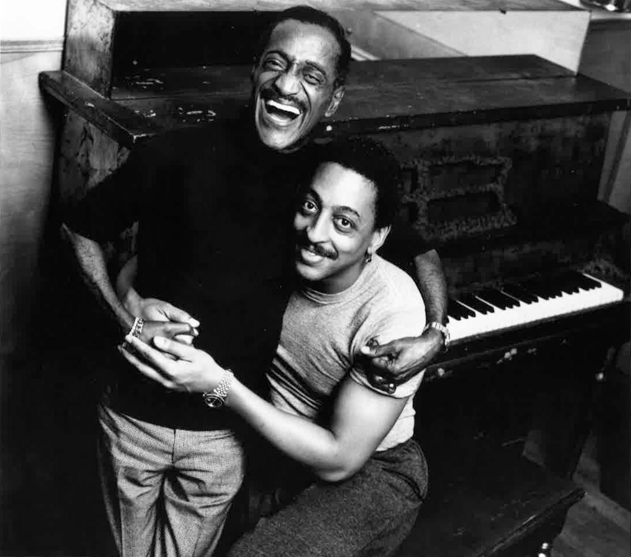 In a black and white archival photo, Sammy Davis Jr. stands beside Gregory Hines, who is seated at a piano bench. Davis Jr. laugh and Hines smiles as the two embrace.