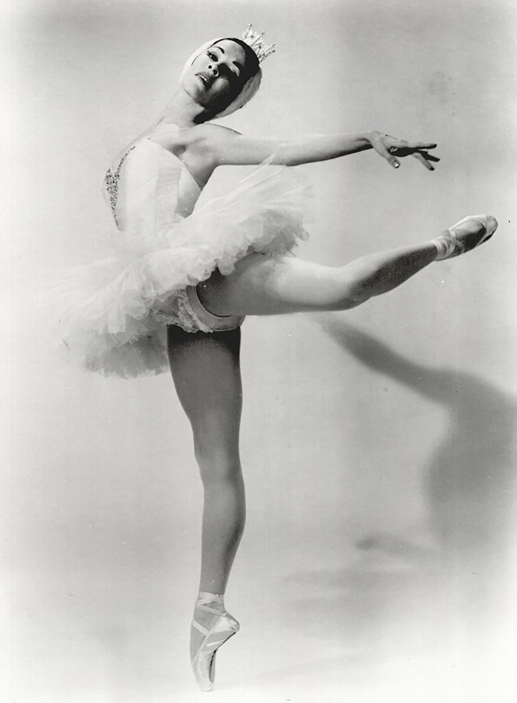 Black and white image of a woman standing in attitude back on pointe, her arms reaching toward her back foot and head angled back