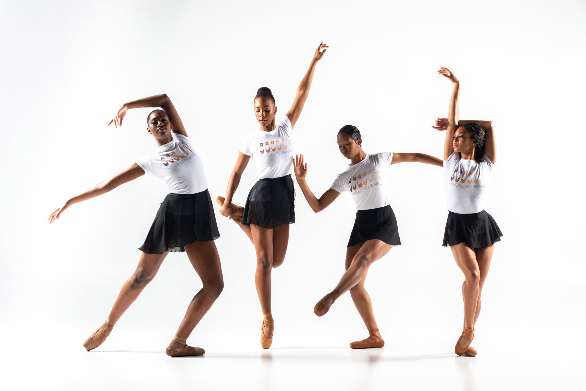 Four Black women pose on a white backdrop wearing pointe shoes that match their varied skin tones, black wrap skirts, and white t-shirts with the Révolutionnaire logo.