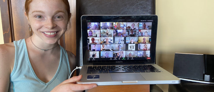 A redhead woman holds up a laptop with the screen showing a Zoom of many screens
