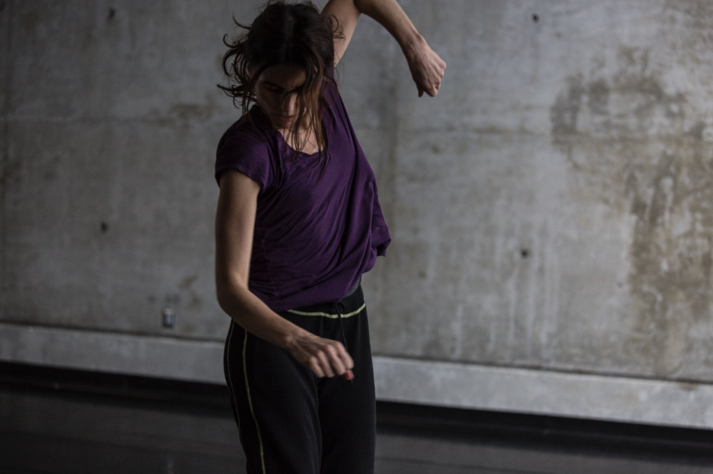 Close-up of Jimena Paz dancing alone in a concrete-walled studio. Her arms loosely near her sides, one up the other down, looking toward the floor