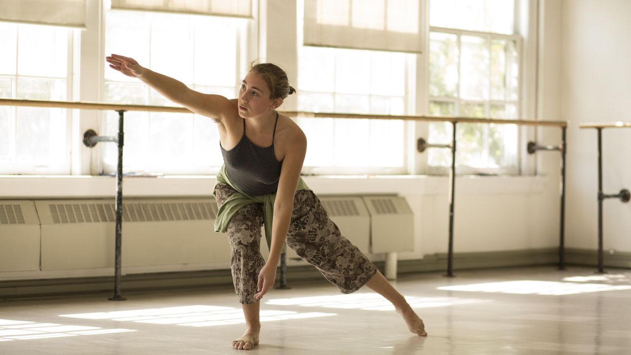 In a sun-drenched studio, a barefoot dancer shallowly lunges with her leg extended side. Her opposite arm is extended to shoulder height as she leans forward, her other arm dangling down.