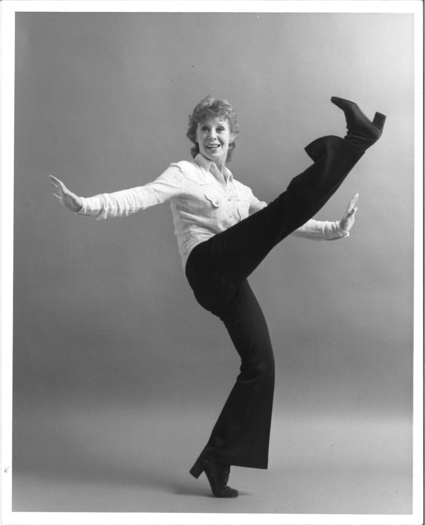In a black and white photo, an older Gwen Verdon smiles charmingly at the camera as she extends her leg in front of her to nose height while balanced on forced arch. Her arms are extended side with flexed hands. She wears a white button down, black pants, and black heeled boots.