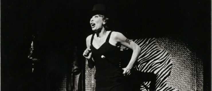 In a black and white archival image, Gwen Verdon sings in a black dress and hat while spotlit, standing atop a bed with a shag white blanket and patterned pillows.
