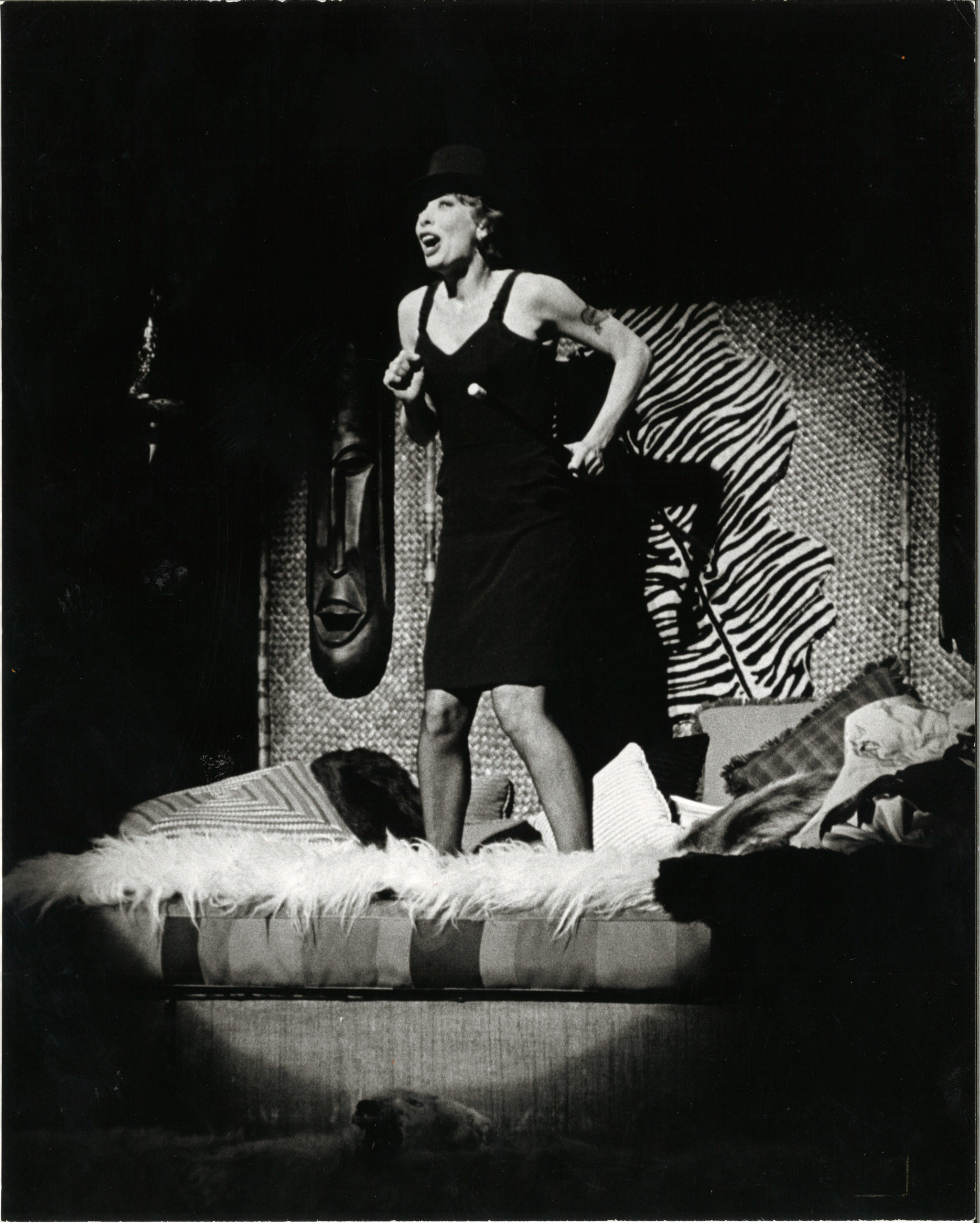 In a black and white archival image, Gwen Verdon sings in a black dress and hat while spotlit, standing atop a bed with a shag white blanket and patterned pillows.