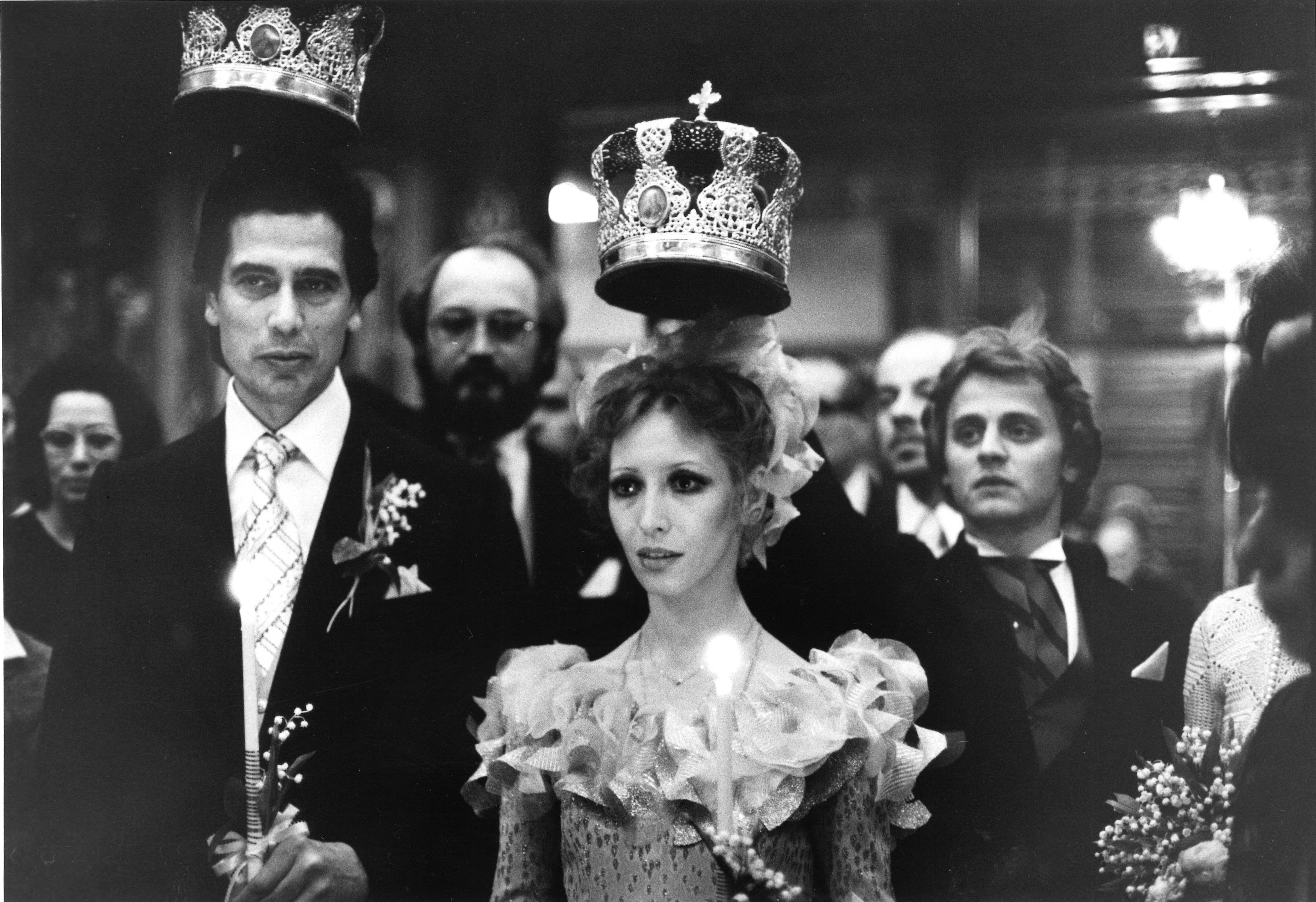 Russian-style crowns are held above Karkar and Makarova's heads, in the latter's case. by Mikhail Baryshnikov. The men wear dark, formal suits, Makarova a dress with a frilled collar.