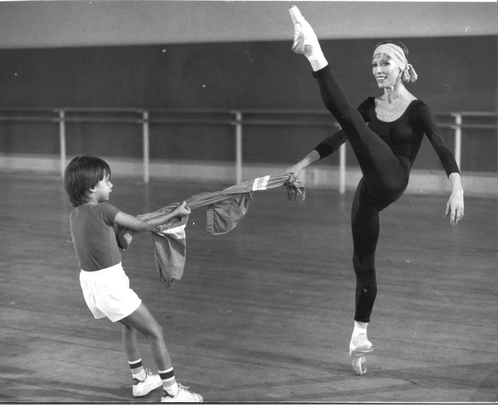 Dressed in all black practice clothes, Makarova smiles widely as she balances en pointe with a leg extended front, the opposite arms holding the end of a sweater being pulled taut by her young son.