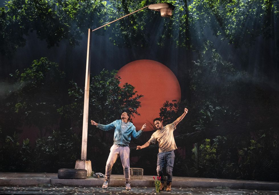 Two Black men dance with their arms up on a stage in front a set of trees and a lamp post.