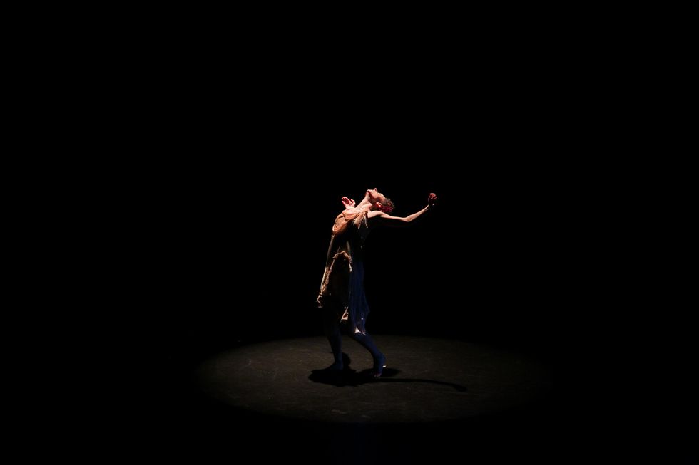 On a dark stage, a spotlight from above illuminates Meredith Fages, arching back with her arms wide and curved at her shoulder height, head thrown back.
