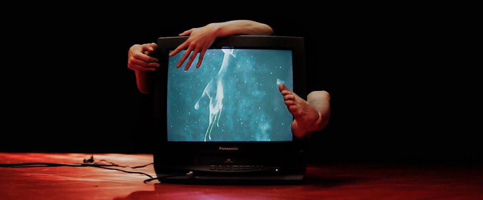 Two hands and a bare foot wrap around a TV showing an image of a hand in water.