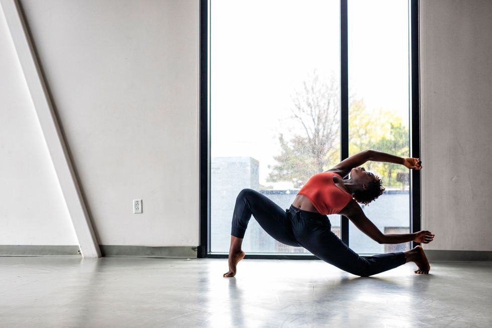 A Black dancer lunges on one knee, feet in forced arch, leaning back with her hands above her heads in a studio in front of a window.