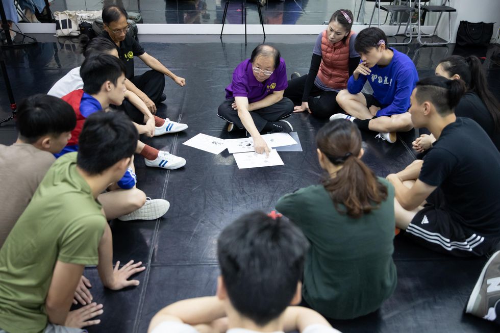 About 12 dancer/researchers sit in a semi circle on the floor of a dance studio. A martial arts teacher in a purple floor sits at center, directing their attention to papers on the floor.