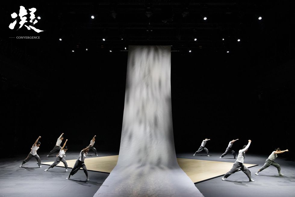 9 dancers perform in "Convergence." They are split into two halves of the stage by a grey cascading stage drop. They all have their right fist clenched above their head and are looking down, in a wide lunge.