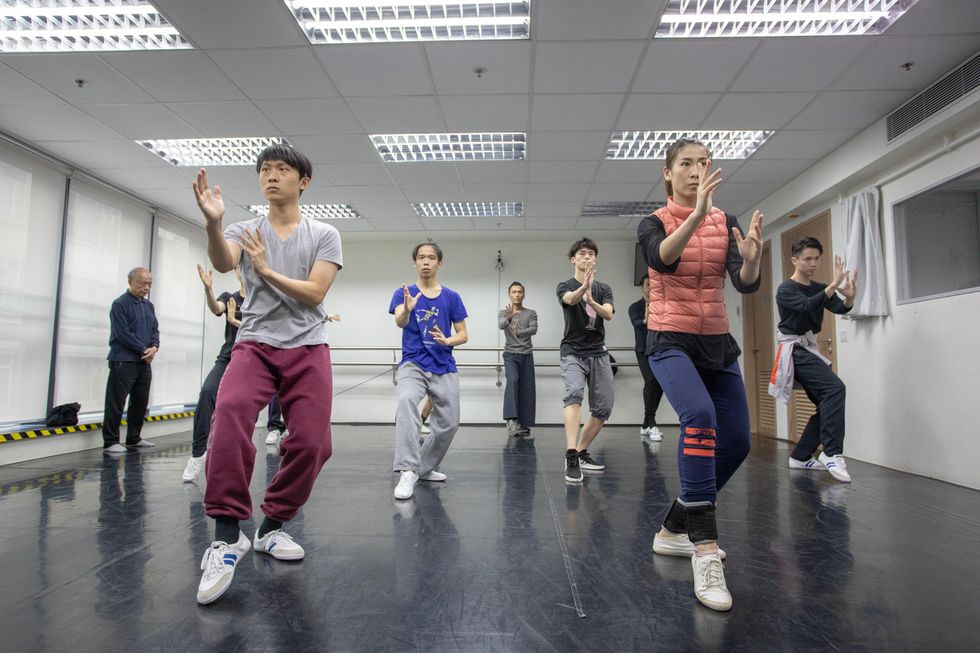 Eight dancer/researchers are in a dance studio for a white crane workshop. They are posed in a pliu00e9 with one leg in front and arms raised in front of them. Their expressions appear very focused.