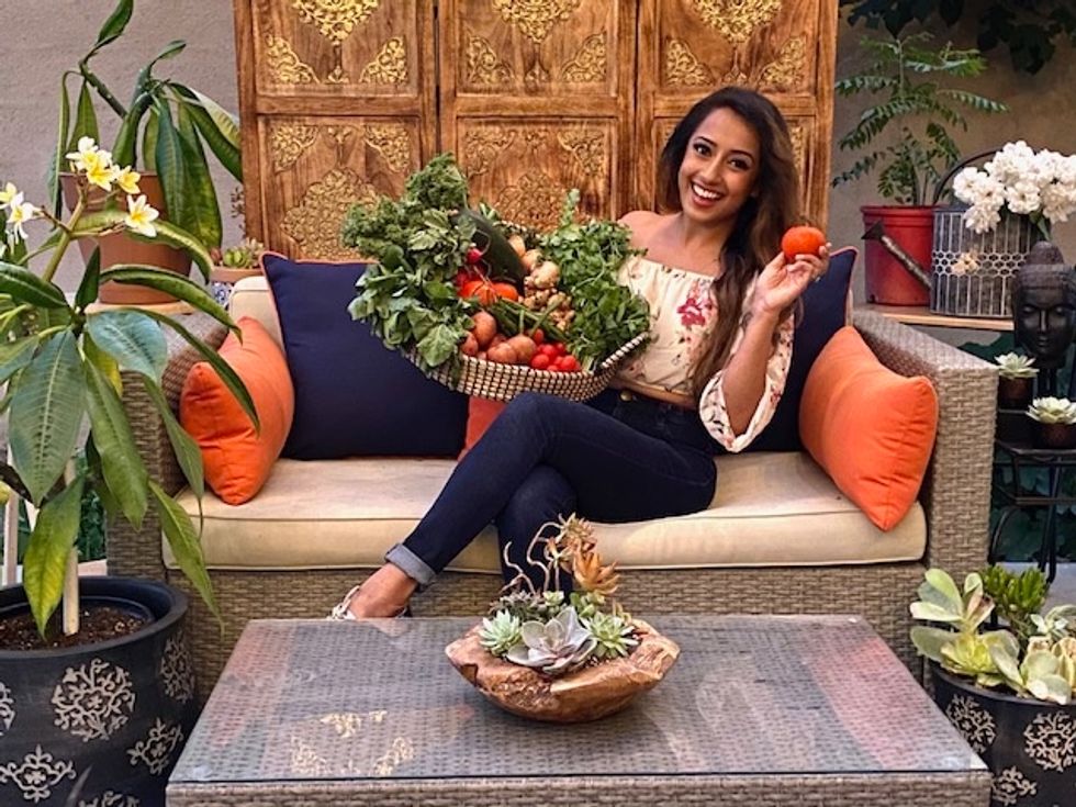 Joya Kazi sits on a couch surrounded by houseplants, holding a large basket of vegetables on her lap and a tomato in one hand.