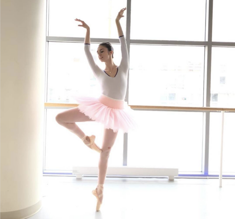 Valle stands on pointe with one leg in passe, arms reaching up and over her head with the backs of her wrists facing each other. She wears a white leotard and pink tutu, and stands in front of floor-to-ceiling windows.