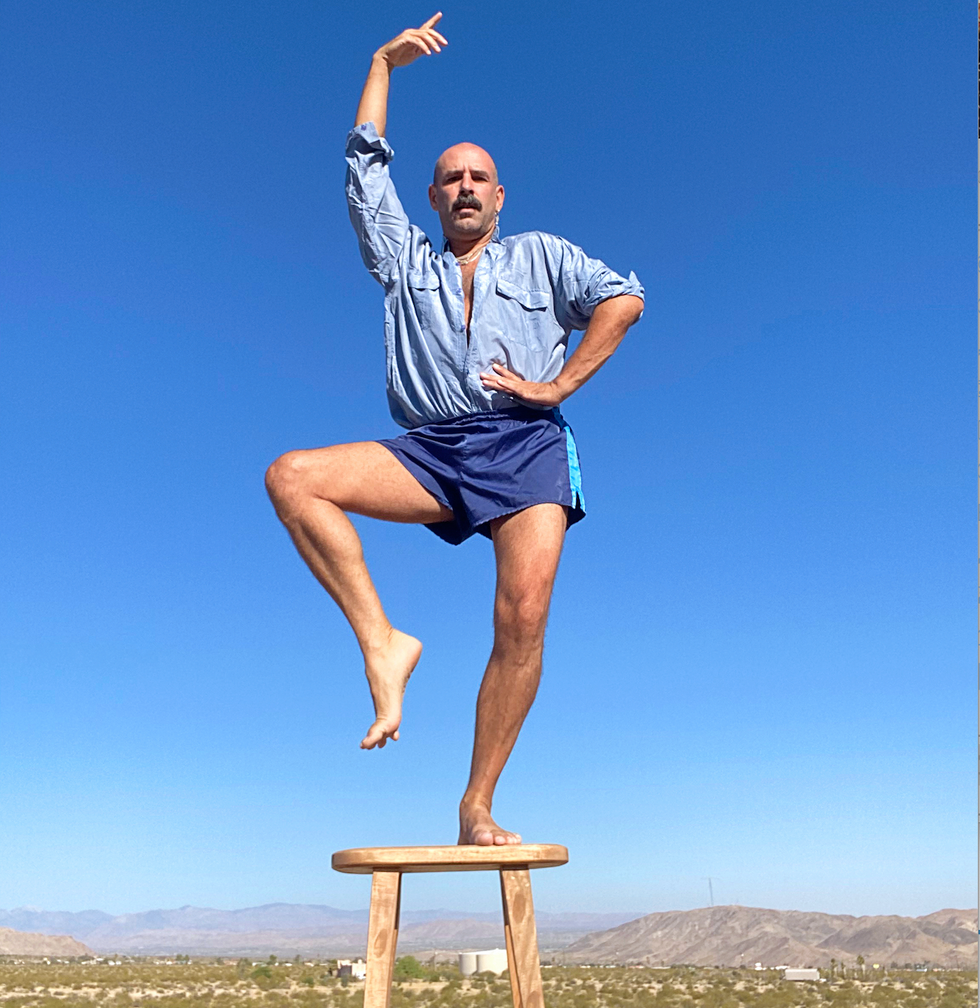 Ryan Heffington stands on a wooden stool in front of a desert landscape, one leg gently raised, toes flexed, hip cocked a bit to the side