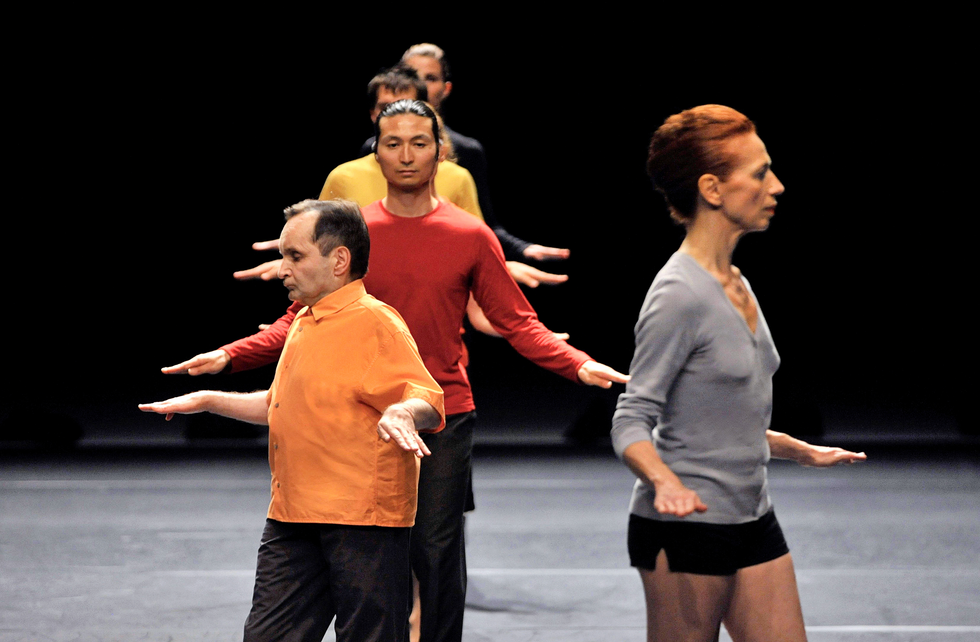 A line of dancers in brightly colored shirts stand with their arms out to the sides