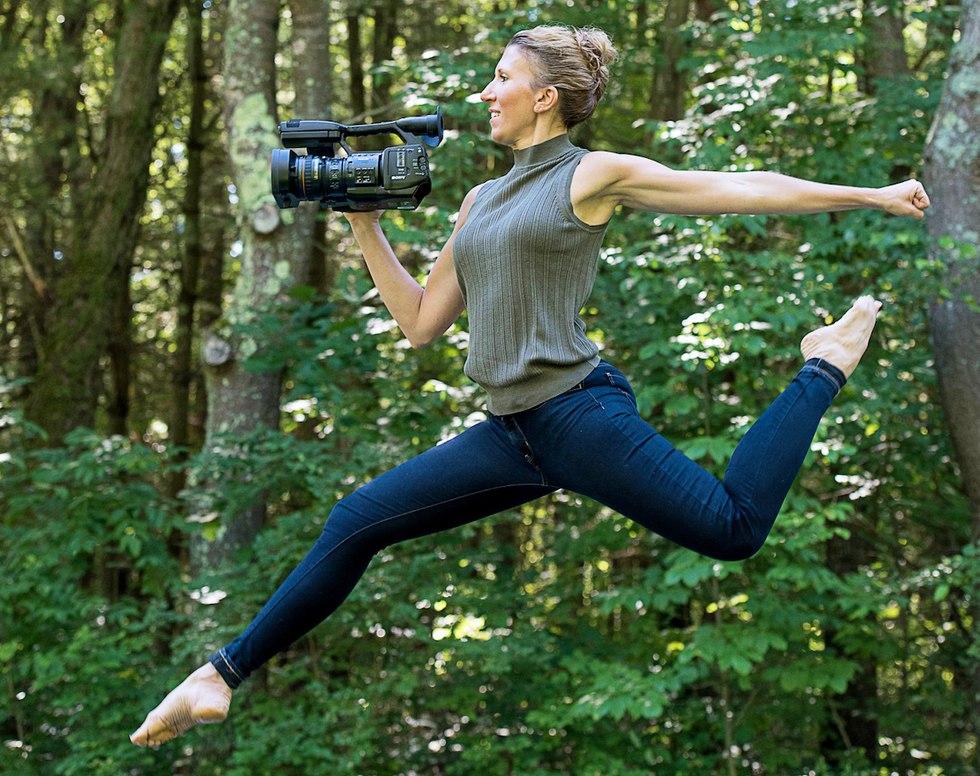 Nel Shelby leaps with a camera in her hand, green trees in the background