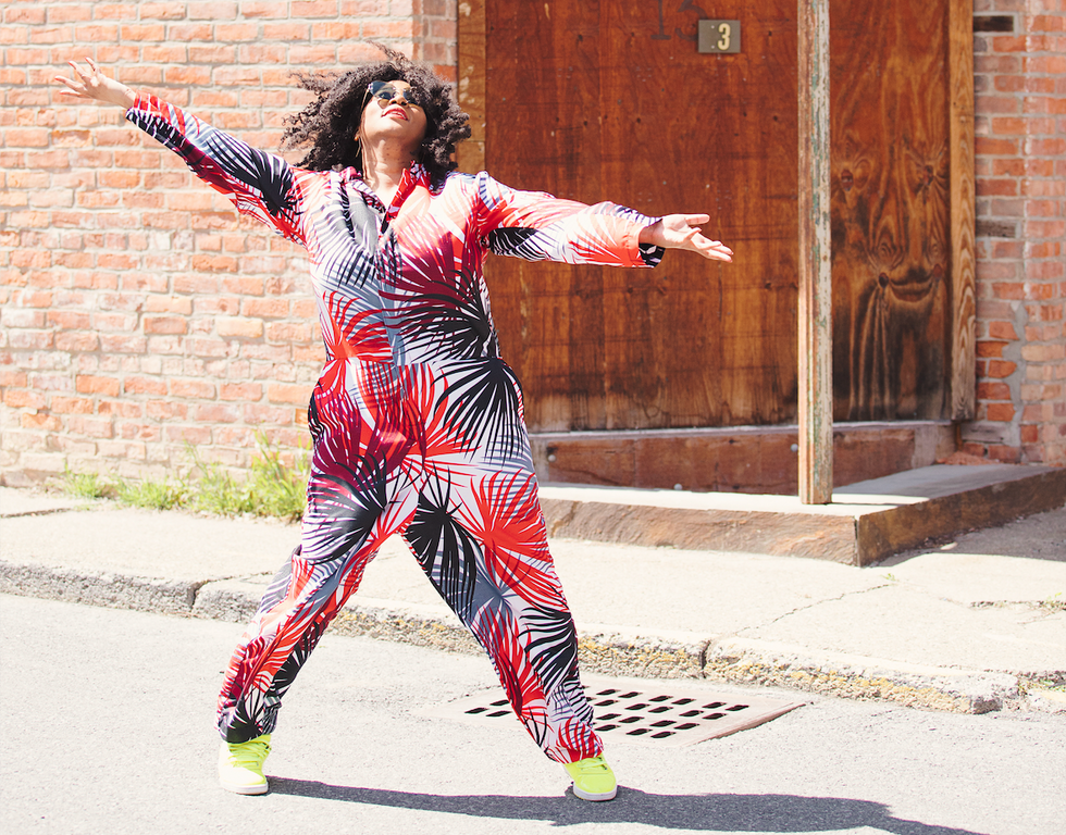 Melanie George in bright pantsuit, stands in wide jazz side lunge on a street in front of a brick building