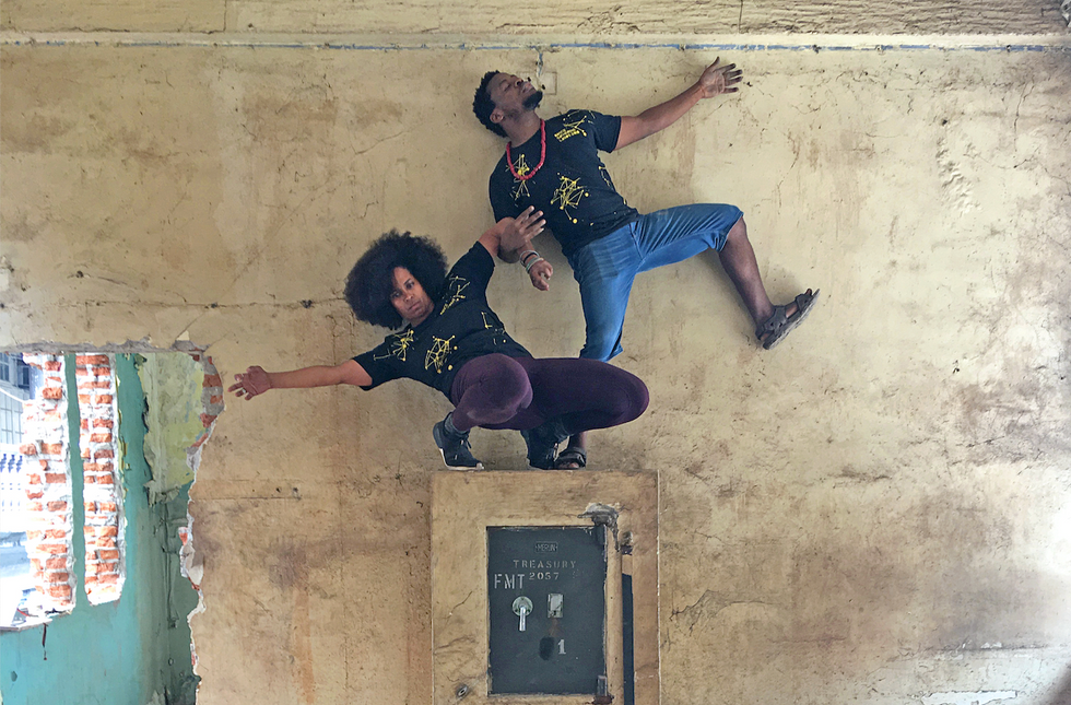 Onye Ozuzu and Qudus Onikeko climb against a paint-cracked wall, on top of what might be a security lock box