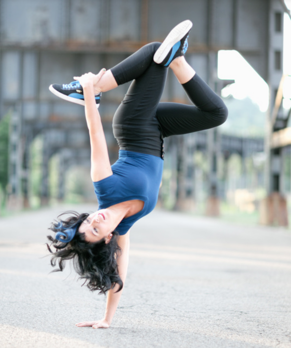 A woman balances on one hand on concrete as her legs fold above her body, her free hand grabbing her back ankle